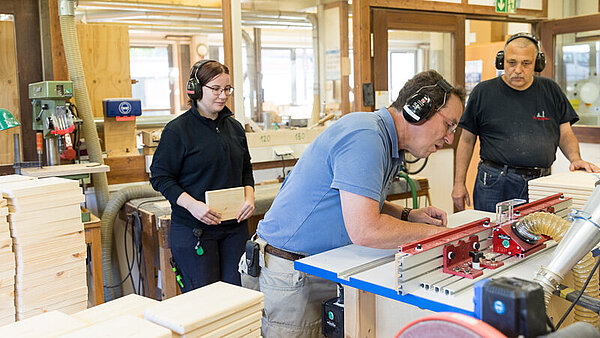 In einer kleinen Werkstatt bearbeitet ein Mann ein Holzstück an einer großen Säge. Zwei weitere Personen stehen daneben und schauen ihm zu.