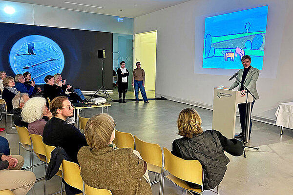 Ein Redner. stehend, spricht vor einem zugewandten Publikum, sitzend, im Ausstellungsraum eines Museums, an den zwei weißen Wänden im Hintergrund sind Kunstprojektionen zu sehen.