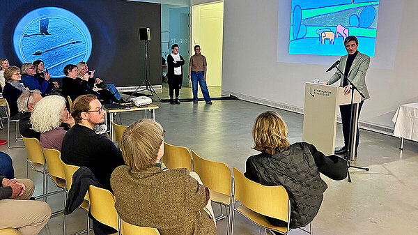 Ein Redner. stehend, spricht vor einem zugewandten Publikum, sitzend, im Ausstellungsraum eines Museums, an den zwei weißen Wänden im Hintergrund sind Kunstprojektionen zu sehen.