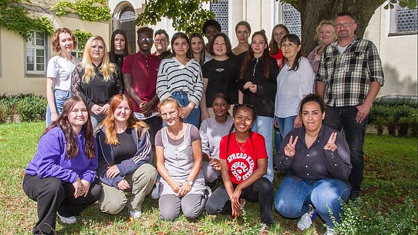 Eine Gruppe von 21 Frauen und Männern steht und sitzt auf einer Wiese unter einem Baum und lacht in die Kamera.