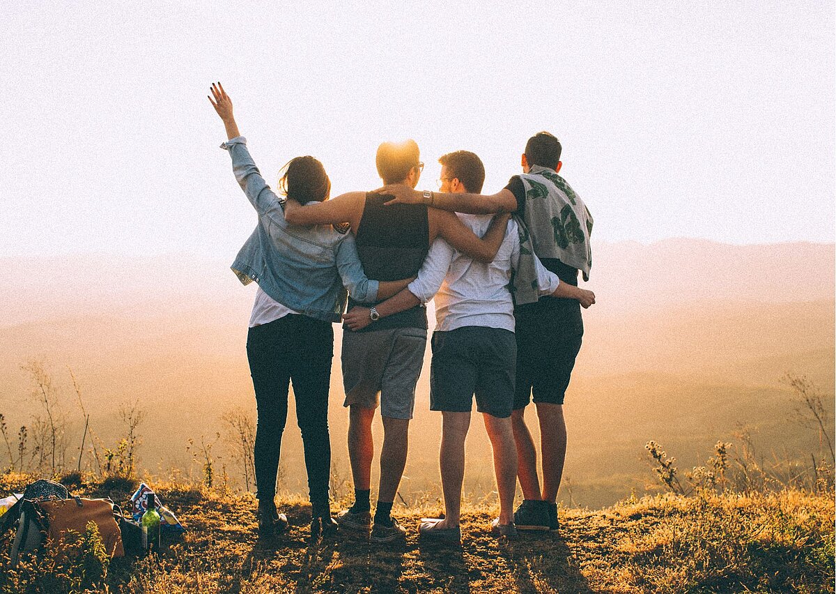 Eine vierköpfige Gruppe junger Leute in Sommerkleidung ist von hinten zu sehen. Sie stehen im Sonnenuntergang auf einer Wiese. 