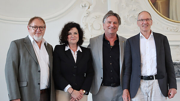 Drei Männer und eine Frau stehen nebeneinander, sie sind alle förmlich gekleidet und lächeln.