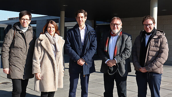 Eine Gruppe von zwei Frauen und drei Männern steht draußen vor einem Gebäude. Die Personen werden von der Sonne angestrahlt und schauen alle in die Kamera.