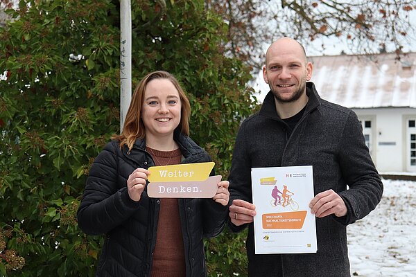 Vor einem Baum stehen eine junge Frau mit mittellangen braunen Haaren (links) und ein Mann mit Bart und Glatze (rechts). Beide halten Papierausdrucke in den Händen und schauen freundlich in die Kamera. 