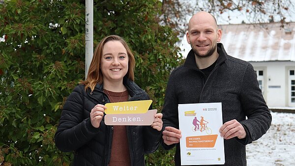 Vor einem Baum stehen eine junge Frau mit mittellangen braunen Haaren (links) und ein Mann mit Bart und Glatze (rechts). Beide halten Papierausdrucke in den Händen und schauen freundlich in die Kamera. 