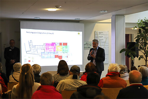 Vor zahlreichen sitzenden Zuhörer:innen steht ein referierender Mann im Sakko neben einer PowerPoint-Präsentation, die einen Grundriss des Krankenhauses Pfullendorf zeigt. 