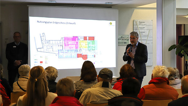 Vor zahlreichen sitzenden Zuhörer:innen steht ein referierender Mann im Sakko neben einer PowerPoint-Präsentation, die einen Grundriss des Krankenhauses Pfullendorf zeigt. 