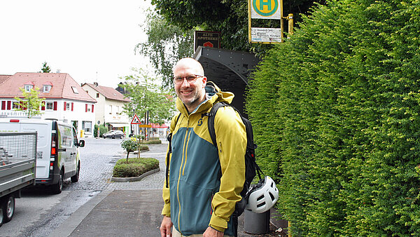 Pressemitteilung Jobticket immer stärker nachgefragt An einer Bushaltestelle an einer Straße steht ein Mann, der freundlich lächelt. Er trägt eine Brille, eine bunte Regenjacke und einen Rucksack, an dem ein Fahrradhelm hängt. Links im Bild fährt ein Transporter mit Anhänger vorbei.