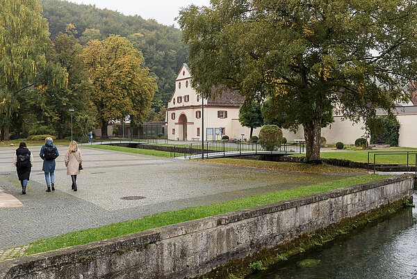Auf einem Platz laufen drei Personen, die von hinten zu sehen sind, nebeneinander spazieren. Im Vordergrund ist ein Kanal, im Hintergrund große Bäume und ein altes Gebäude.