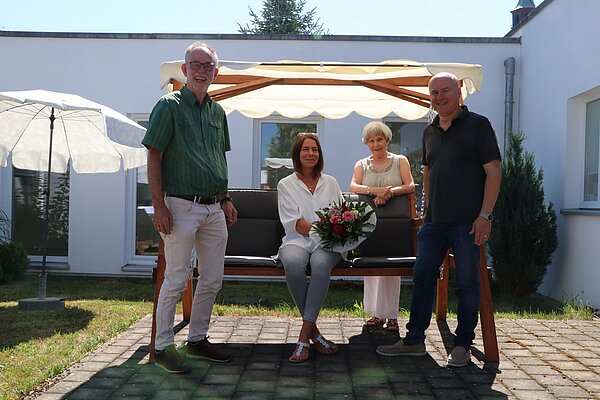 Vier Personen, 2 Frauen und 2 Männer, stehen gut gelaunt in der Sonne vor einer Hollywoodschaukel.