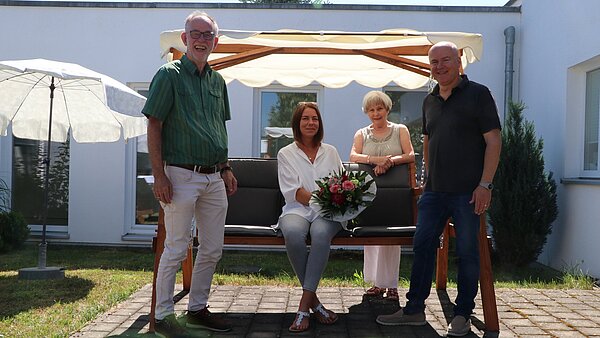 Vier Personen, 2 Frauen und 2 Männer, stehen gut gelaunt in der Sonne vor einer Hollywoodschaukel.
