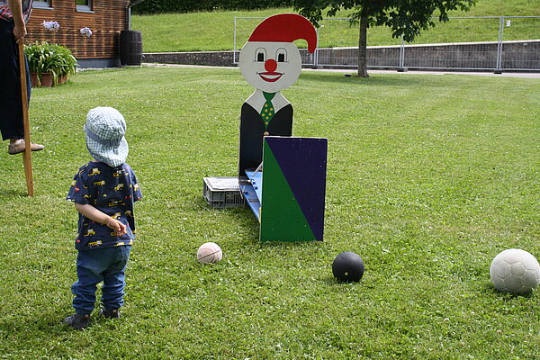 Auf einer grünen Wiese steht eine bunte Holzfigur, davor liegen mehrere Bälle im Gras. Links steht ein kleiner Junge mit Mütze.