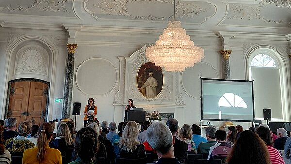 In einem großen Saal mit Stuckdecke und Kronleuchter sitzen die Zuschauer und schauen auf eine Bühne. 