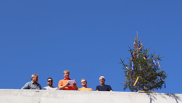 Man sieht 5 Männer, die auf einen Betongebäude neben einem bunt geschmücktem Tannenbaum stehen.