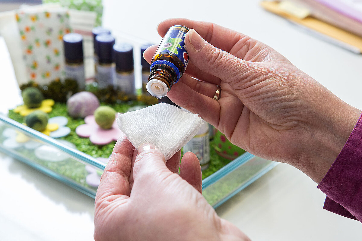 Eine Hand hält links einen Tupfer, die Hand rechts hält ein Glasfläschchen Aromaöl über den Wattetupfer. Im Hintergrund ist ein Tablett mit verschiedenen Sachen wie Glasfläschchen drauf.