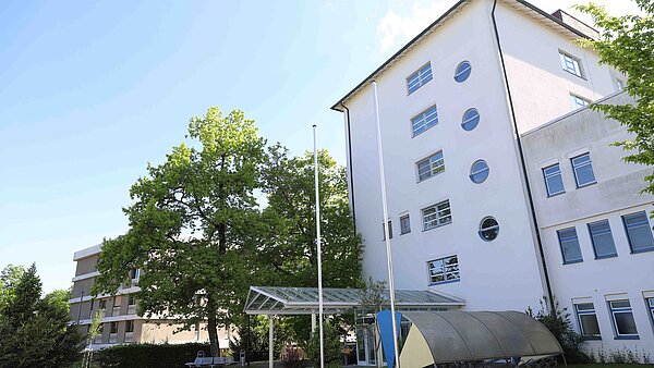 Man sieht ein mehrstöckiges helles Gebäude mit einem überdachten Eingang. 