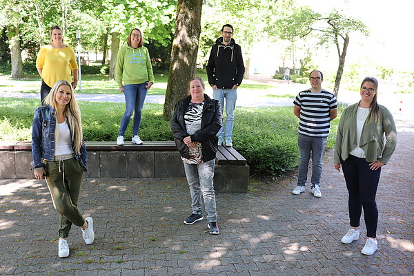 4 Menschen stehen auf dem Boden, dahinter sind 3 Menschen auf einer Bank in einem Park stehen. Alle schauen zur Kamera.