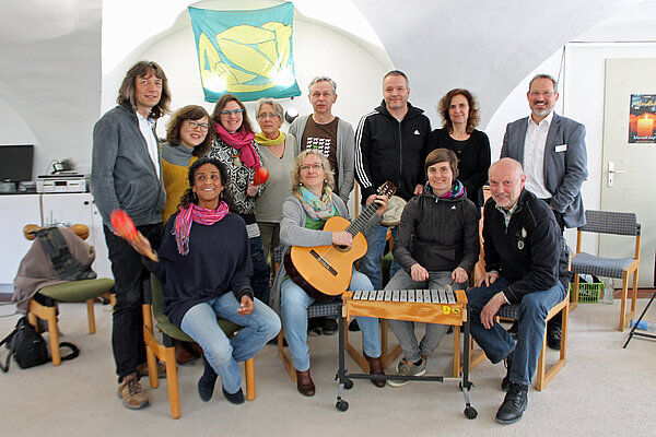Gruppe von Musiktherapeuten stellt sich vor. 12 Menschen sind sitzend und stehend zu sehen. in der Mitte sitzt eine Frau mit Gitarre und im Vordergrund ist ein Xylophon zu sehen.