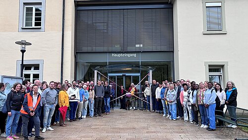 Eine große Gruppe Menschen steht vor einem mehrstöckigen Gebäude, im Hintergrund ist ein Eingang zu erkennen.