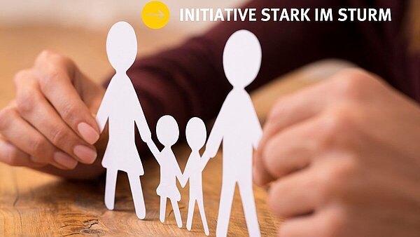 Zu sehen sind zwei Hände auf einem Holztisch, die vier weiße Scherenschnitt-Figuren zeigen, welche Familie symbolisieren sollen.