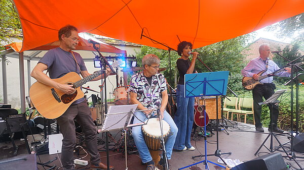 Drei Männer und eine Frau stehen mit ihren Instrumenten auf einer Bühne unter einem orangefarbenen Sonnensegel. 