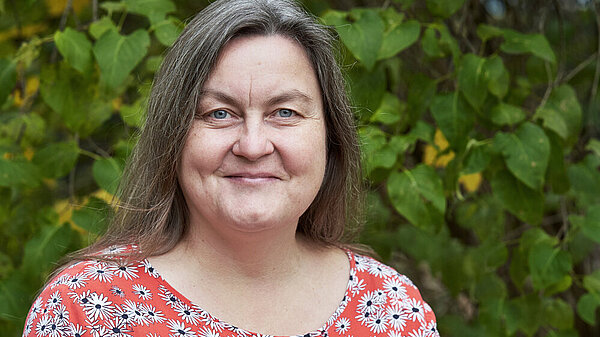 Eine dunkelhaarige Frau mit halblangen Haaren, blauen Augen und einer geblümten Bluse. 