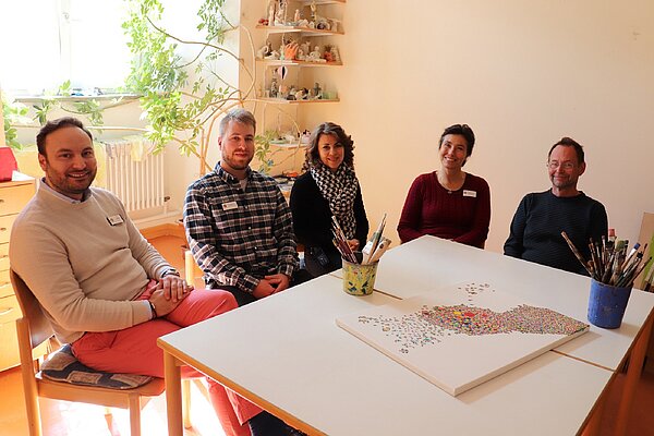 An einem Tisch sitzen nebeneinander drei Männer und zwei Frauen dazwischen und schauen freundlich in die Kamera.