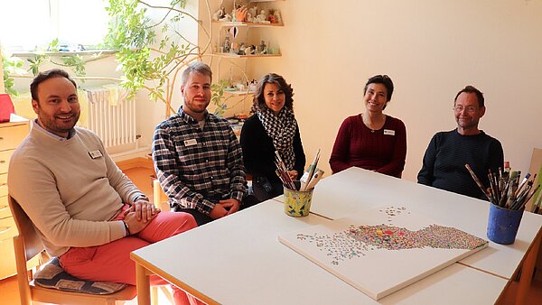 An einem Tisch sitzen nebeneinander drei Männer und zwei Frauen dazwischen und schauen freundlich in die Kamera.