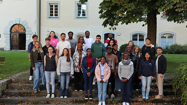 Gruppenfoto von 20 Personen, die auf einer Treppe vor einem Gebäude stehen. Die meisten tragen Freizeitkleidung, einige mit Jacken und Pullovern. Bäume im Hintergrund.
