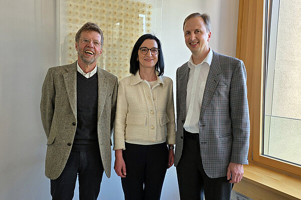 Eine Frau und zwei Männer stehen in einem Büroraum nebeneinander und lächeln in die Kamera.