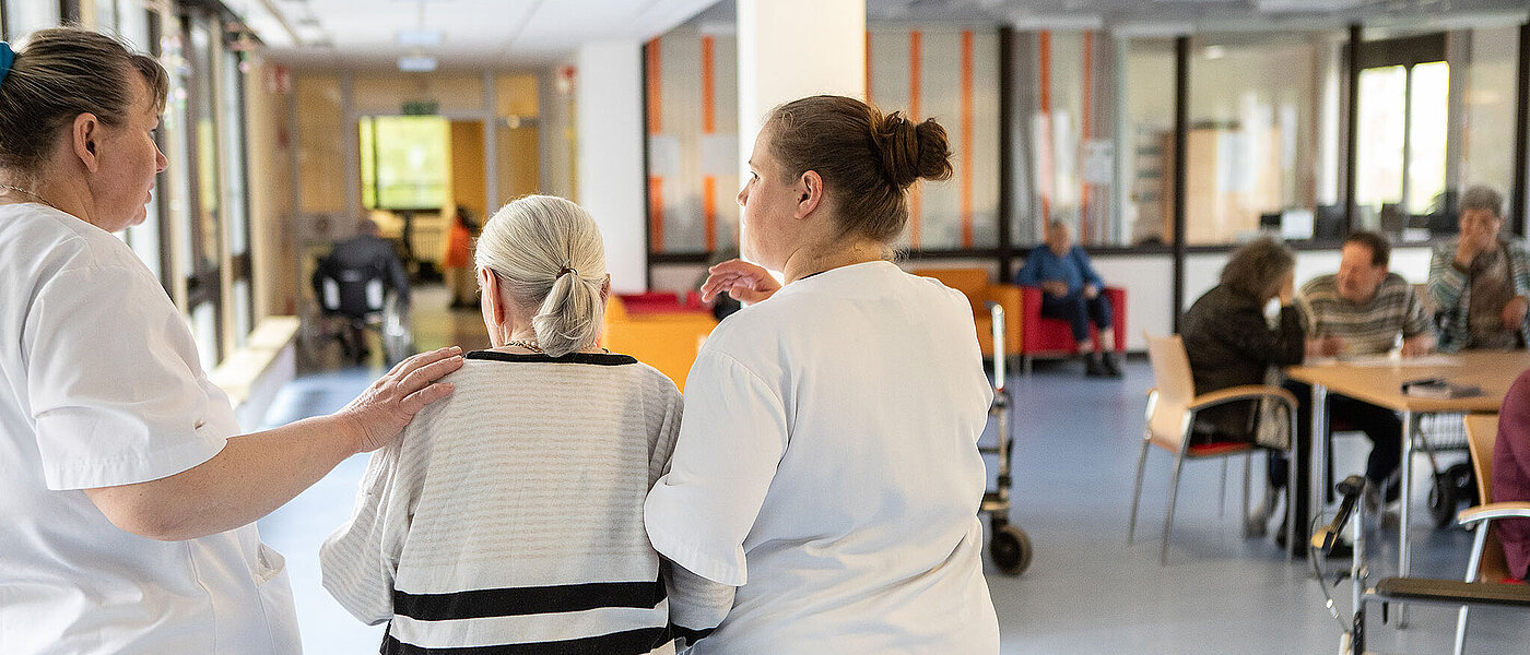 Eine junge Pflegerin unterstützt eine ältere Dame im blau gestreiften Pullover beim Gehen