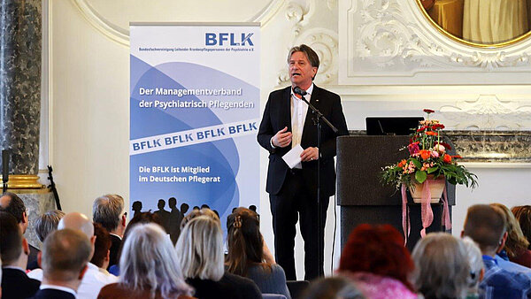 Ein Mann in weißem Hemd und schwarzem Anzug steht in einem historischen Festsaal mit stuckverzierten Wänden vor zahlreichen Zuhöherinnen und Zuhörern.