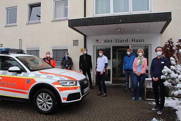 Ein Rettungsarzt Fahrzeug und sieben Personen stehen vor dem Abt-Siard Haus Eingang nebeneinander in Abstand und FFP2 Maske.