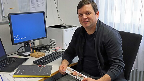 Ein Mann, Marian Jurcsik, sitzt auf einem Stuhl an einem Schreibtisch. In der Hand hält er den Newsletter von Beclean e.V..