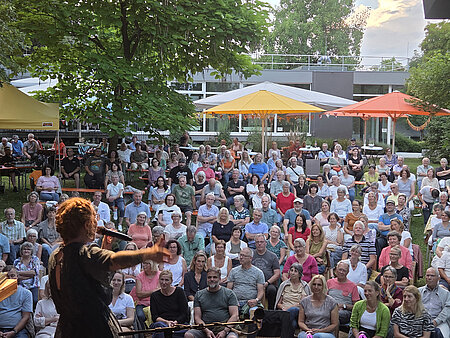 Ber Blick von hinter der Bühne zeigt eine Künstlerin, die vor einer großen Menschenmenge bei einer Open-Air-Veranstaltung steht.