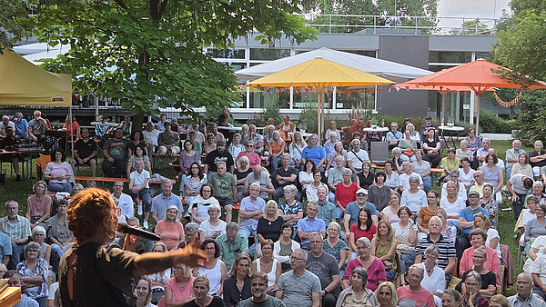 Ber Blick von hinter der Bühne zeigt eine Künstlerin, die vor einer großen Menschenmenge bei einer Open-Air-Veranstaltung steht.