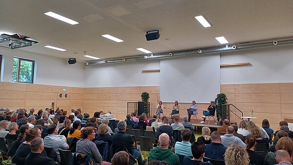 In einem großen Saal sitzen viele Menschen nebeneinander auf Stühlen und blicken konzentriert zur Bühne, auf der vier Frauen nebeneinander sitzen und vortragen. 