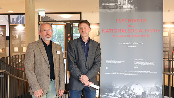 Zwei Personen stehen neben einem Informationsbanner zur Wanderausstellung 'Psychiatrie und Nationalsozialismus im deutschen Südwesten 1933-1945'. Das Banner zeigt historische Fotos und Text.