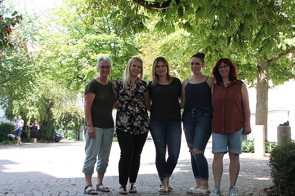 fünf Damen stehen eng beinander und haben die Arme um sich gelegt. 