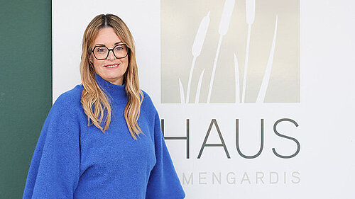 Eine blonde Frau im blauen Pullover steht auf einem Schild auf dem "Haus Irmengardis" steht.