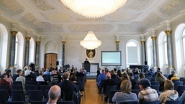 Blick in einen barocken Festssaal mit Holzparkett, Stuck und Kronleuchtern, in dem zahlreiche Zuhörer:innen auf Stuhlreihen einem Redner am Rednerpult gegenüber sitzen.