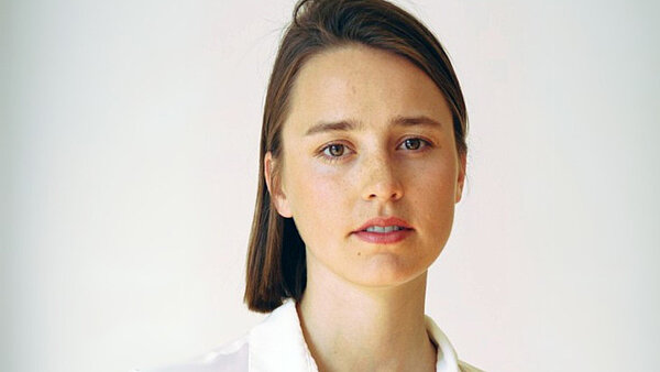 Eine junge Frau mit brünettem, glatten Haar und weißer Bluse. 