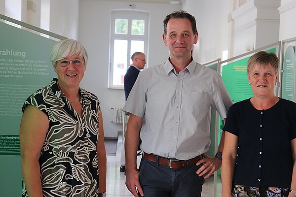 Ein Mann steht zwischen zwei Damen, im Hintergrund sind grüne Stelen einer Ausstellung zu erkennen. 