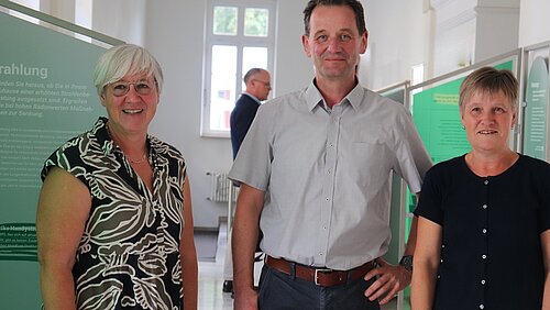Ein Mann steht zwischen zwei Damen, im Hintergrund sind grüne Stelen einer Ausstellung zu erkennen. 