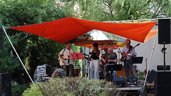 Vier Musiker auf einer kleinen Bühne im Freien unter einem orangen Sonnensegel, umgeben von Büschen und Bäumen.