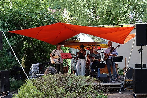 Vier Musiker auf einer kleinen Bühne im Freien unter einem orangen Sonnensegel, umgeben von Büschen und Bäumen.
