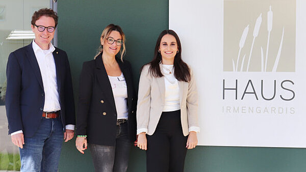 Ein Mann und zwei Frauen stehen vor einem Schild mit der Aufschrift "Haus Irmengardis". 