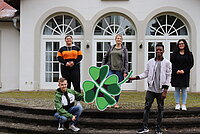 Fünf junge Menschen stehen nebeneinander vor einem Gebäude, zwei von ihnen halten ein überdimensionales Kleeblatt in den Händen. 