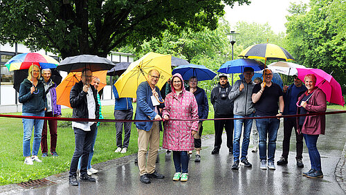 Ein Mann und eine Frau stehen im Vordergrund und durchschneiden symbolisch ein Seil, im Hintergrund stehen Zuschauende; alle haben sie Regenschirme über sich, da es in Strömen regnet.
