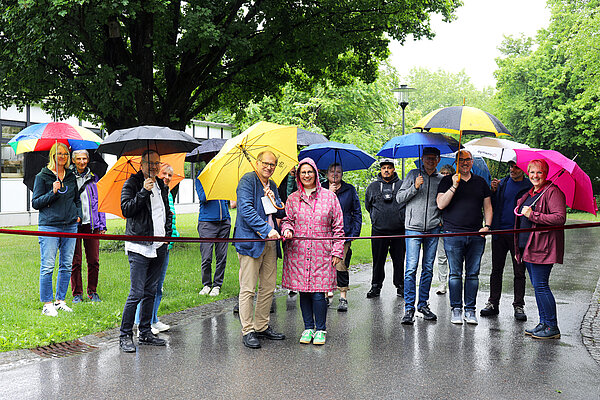 Ein Mann und eine Frau stehen im Vordergrund und durchschneiden symbolisch ein Seil, im Hintergrund stehen Zuschauende; alle haben sie Regenschirme über sich, da es in Strömen regnet.
